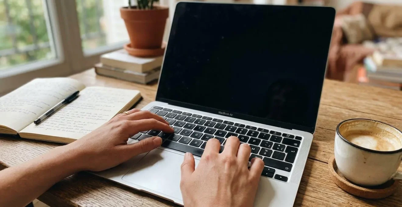 Gros plan sur des mains tapant sur un clavier d'ordinateur portable, avec un carnet de notes et une tasse de café posés sur un bureau en bois clair